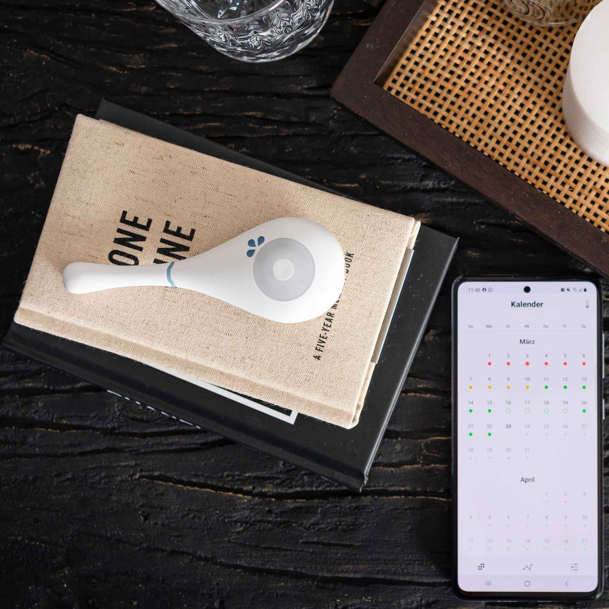 Daysy fertility tracker resting on a cloth-bound book beside a smartphone displaying a calendar app, on a wooden nightstand