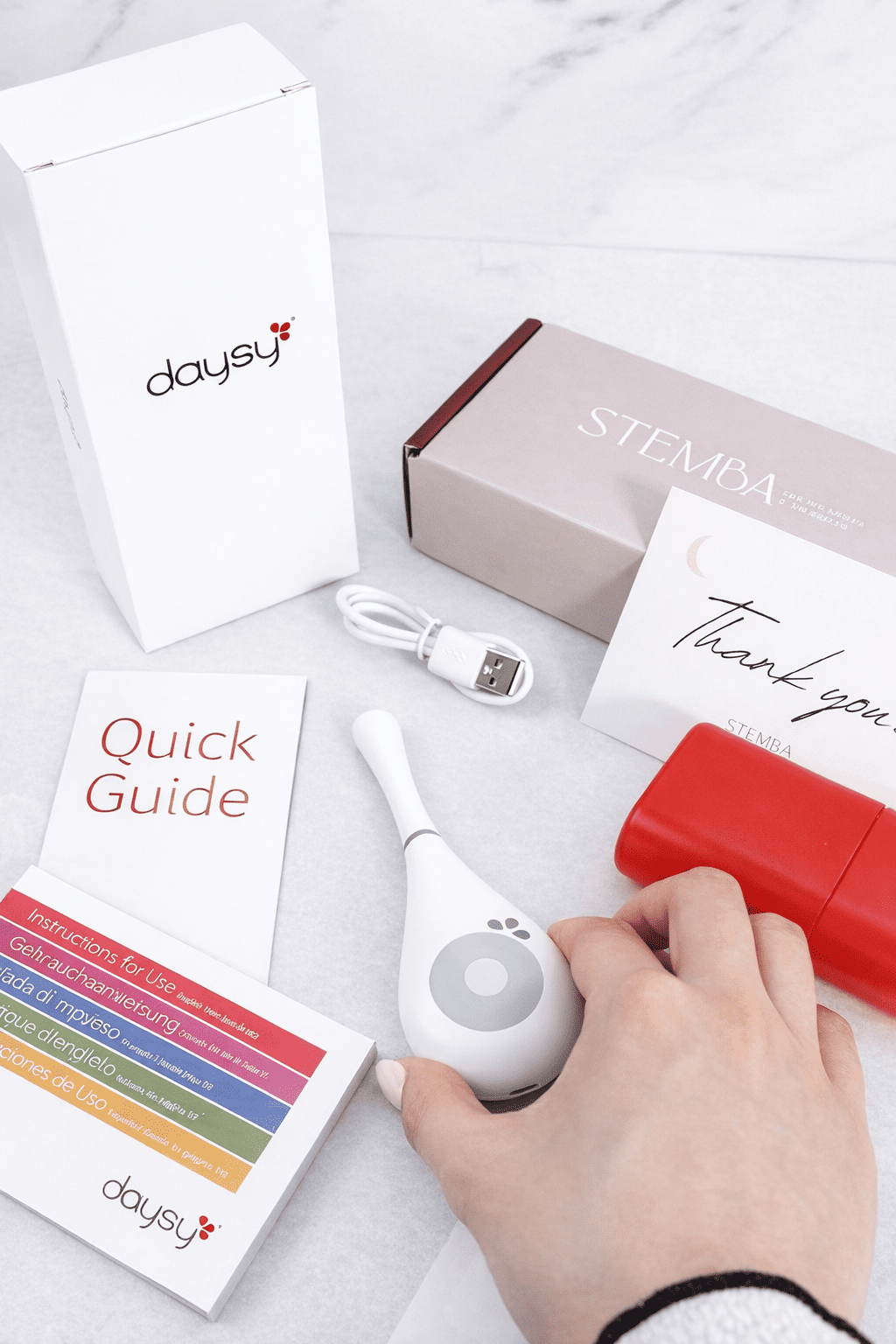 Daysy fertility tracker unboxing flatlay — device, red storage case, USB cable, quick guide and Stemba packaging on marble surface