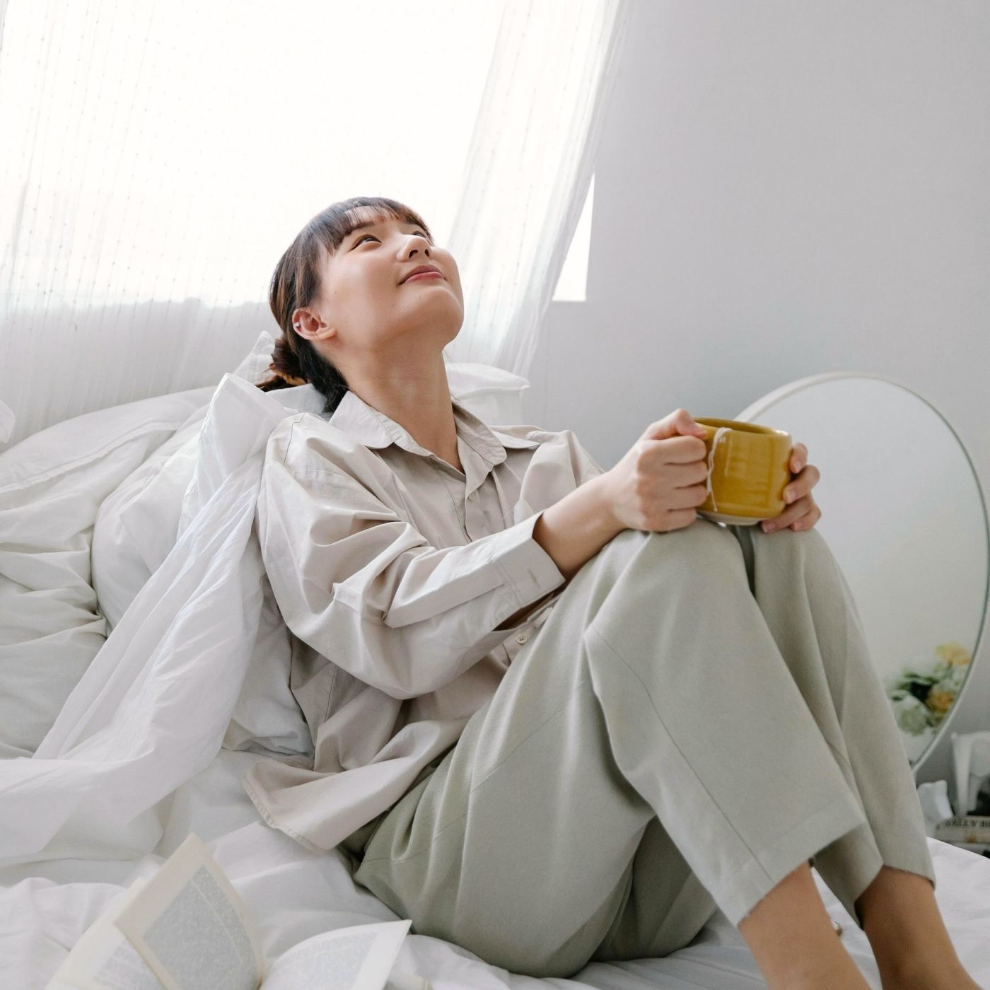 Woman sitting on bed holding a cup of coffee and looking relaxed in soft morning light